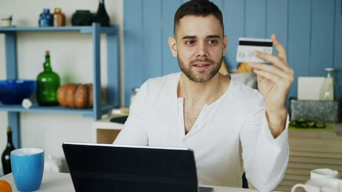 A smiling man at a laptop holds up a credit card while applying for credit online at home