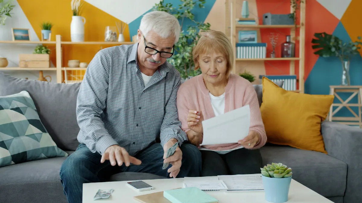 An older couple sits on a sofa reviewing paperwork and bills at a coffee table