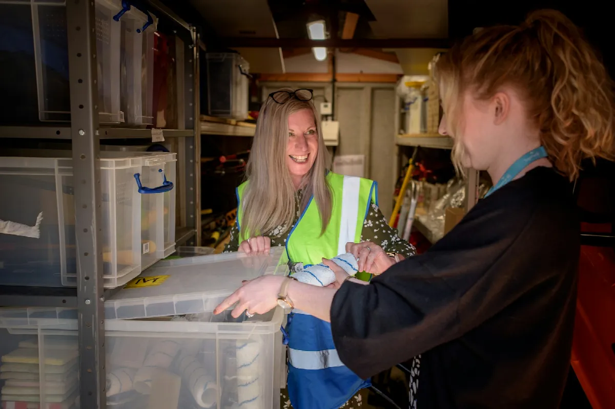 A woman in a safety vest and a younger coworker check a folder of paperwork in a stockroom lined with storage bins