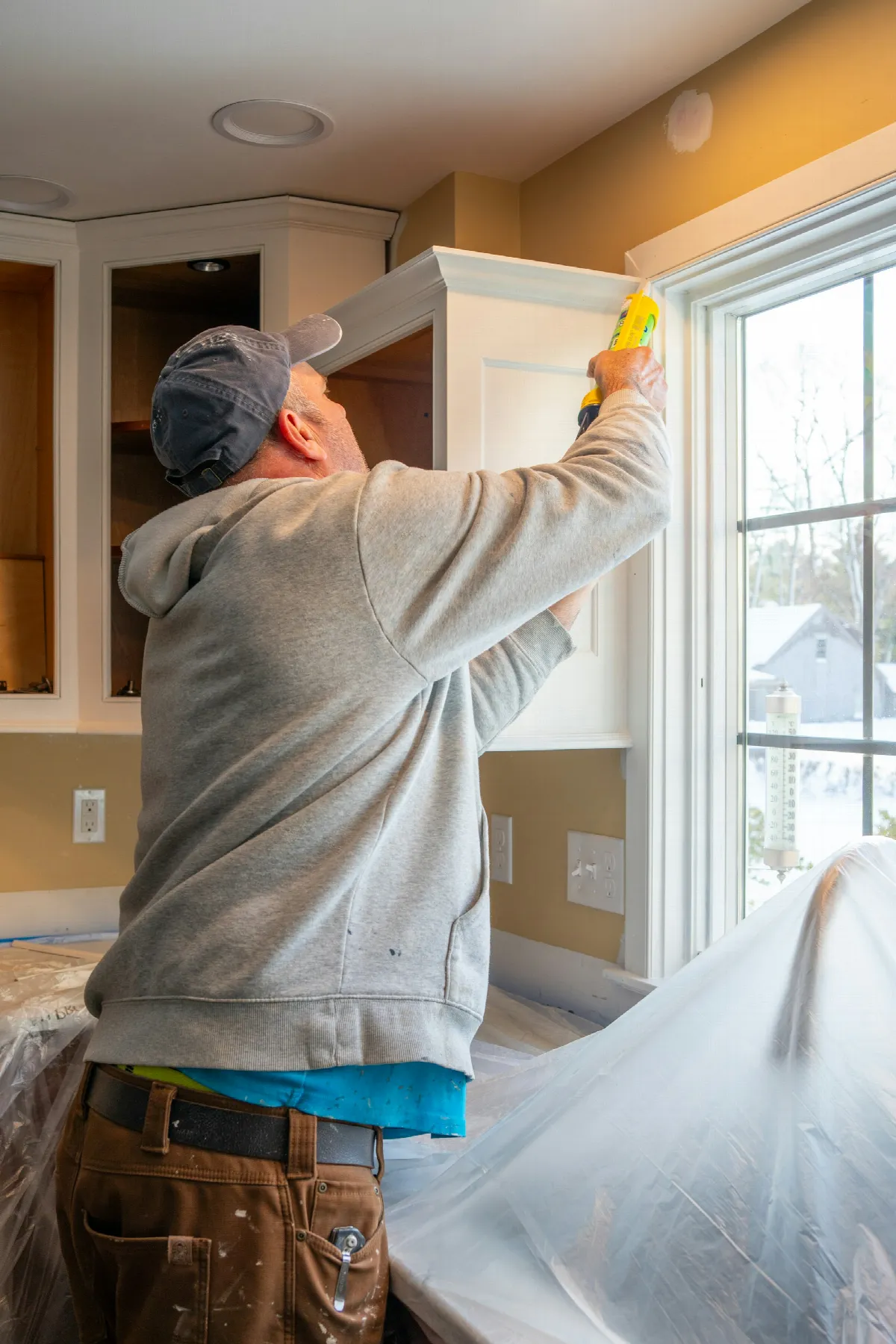 A contractor in a gray hoodie caulks the trim around a kitchen window while the counters are covered in plastic sheeting