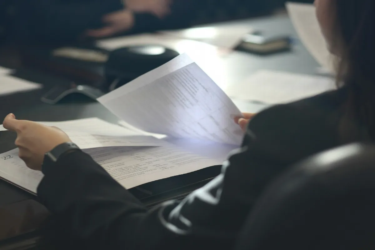A person at a dark desk holding up a printed page while reading through a stack of documents
