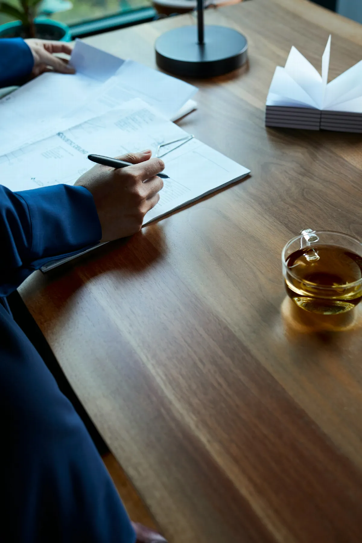 A person in a dark blazer reviewing a printed document with a pen at a wooden desk, a cup of tea beside them