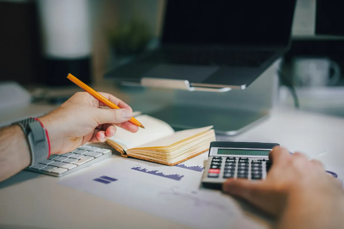 A person working out card balances on a calculator and in a notebook to figure their credit utilization, a laptop open behind them
