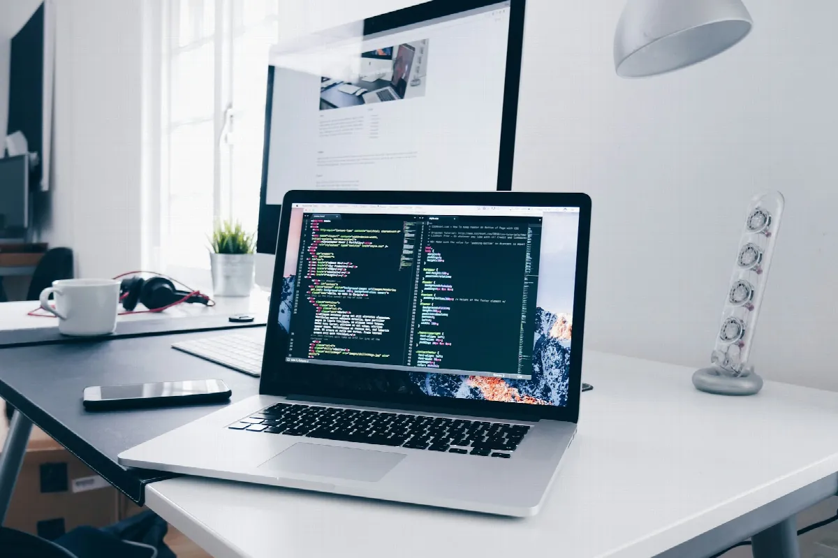 A laptop showing lines of code on a bright white desk next to a monitor, a lamp and a phone