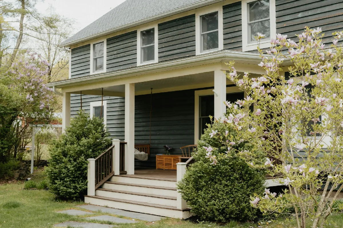 A gray two-story house with a covered front porch, white railings and flowering shrubs by the steps
