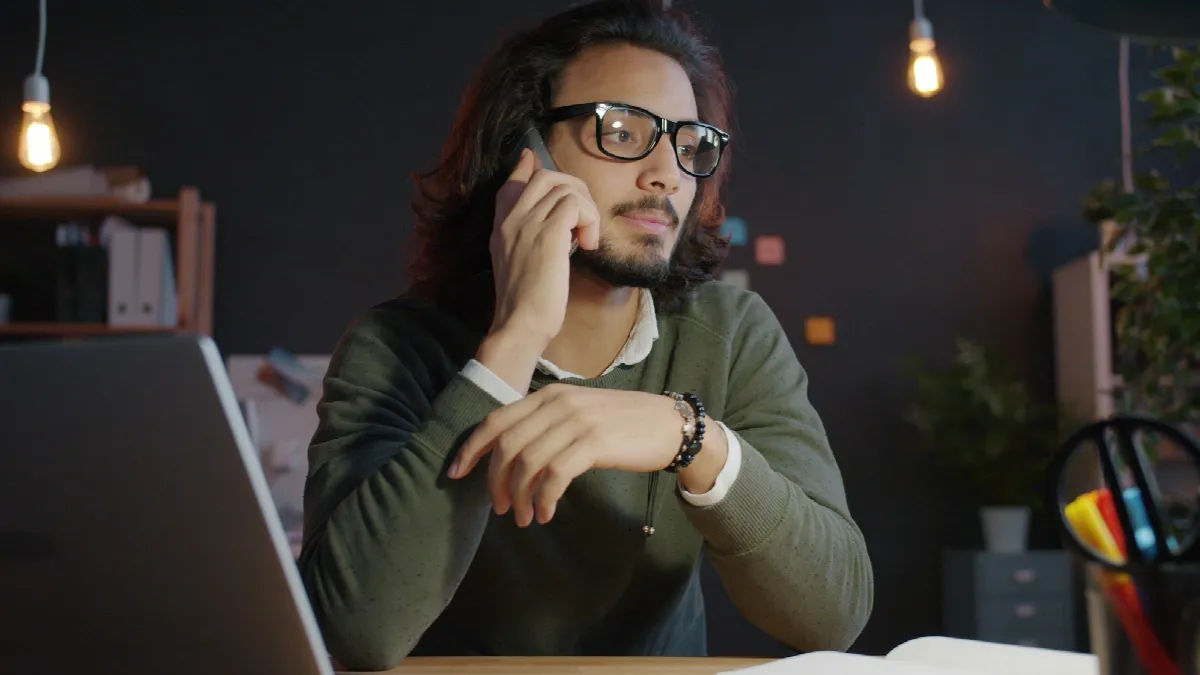 A man in glasses on a phone call at his desk, looking off to the side as if trying to place who's calling