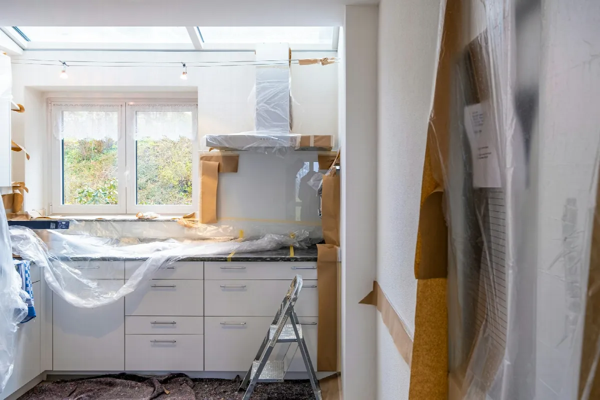 A kitchen mid-renovation, with white cabinets covered in plastic sheeting and brown paper on the floor