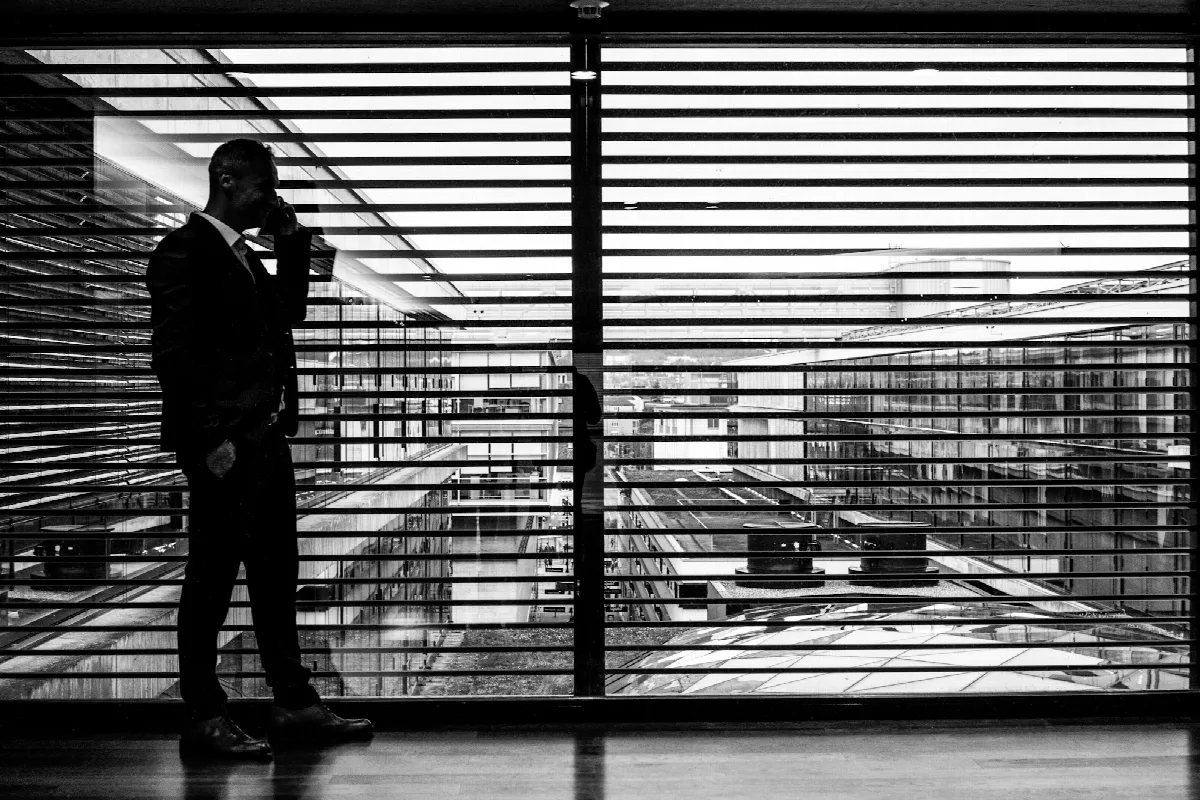 Silhouette of a man on a phone call standing beside a large window with horizontal blinds