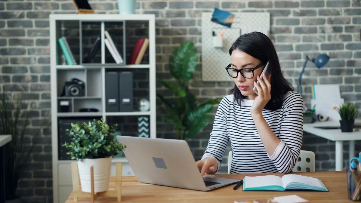 A woman in glasses talks on her phone while typing on a laptop at a desk in a bright office