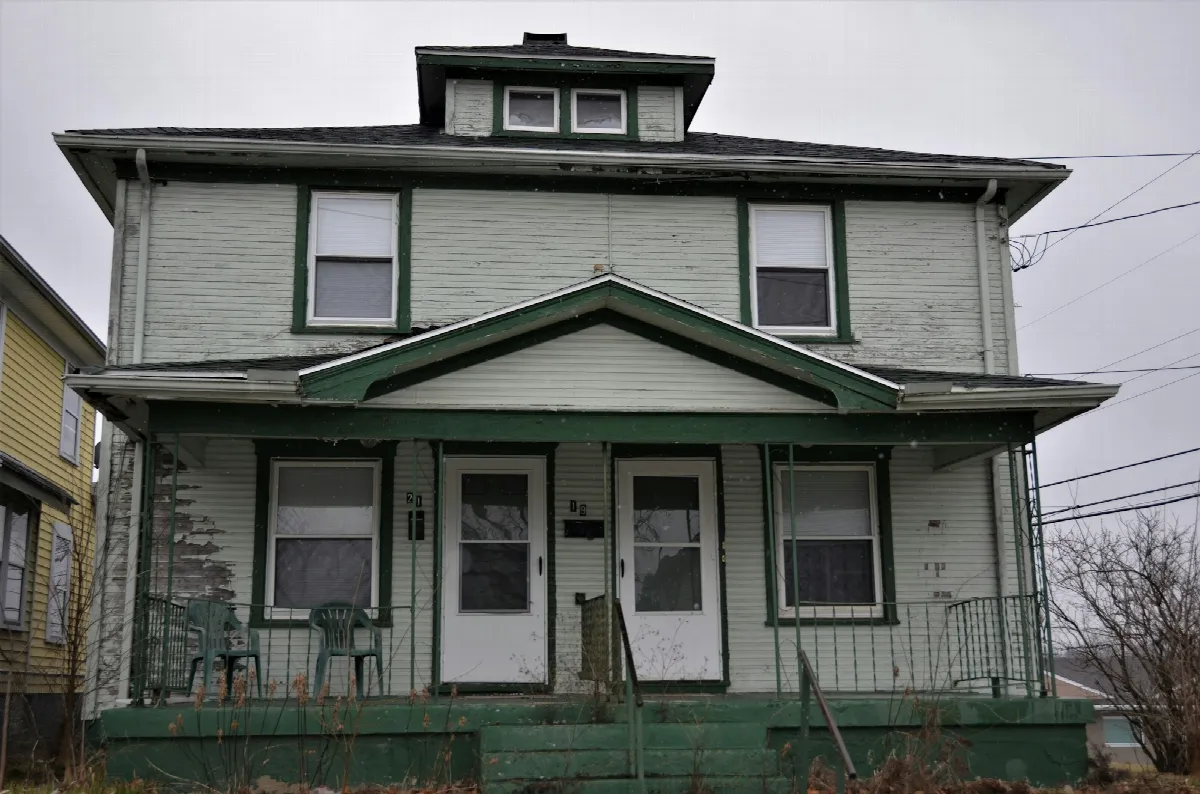 A two-story duplex with two front doors, a shared green porch roof and bare trees on an overcast day