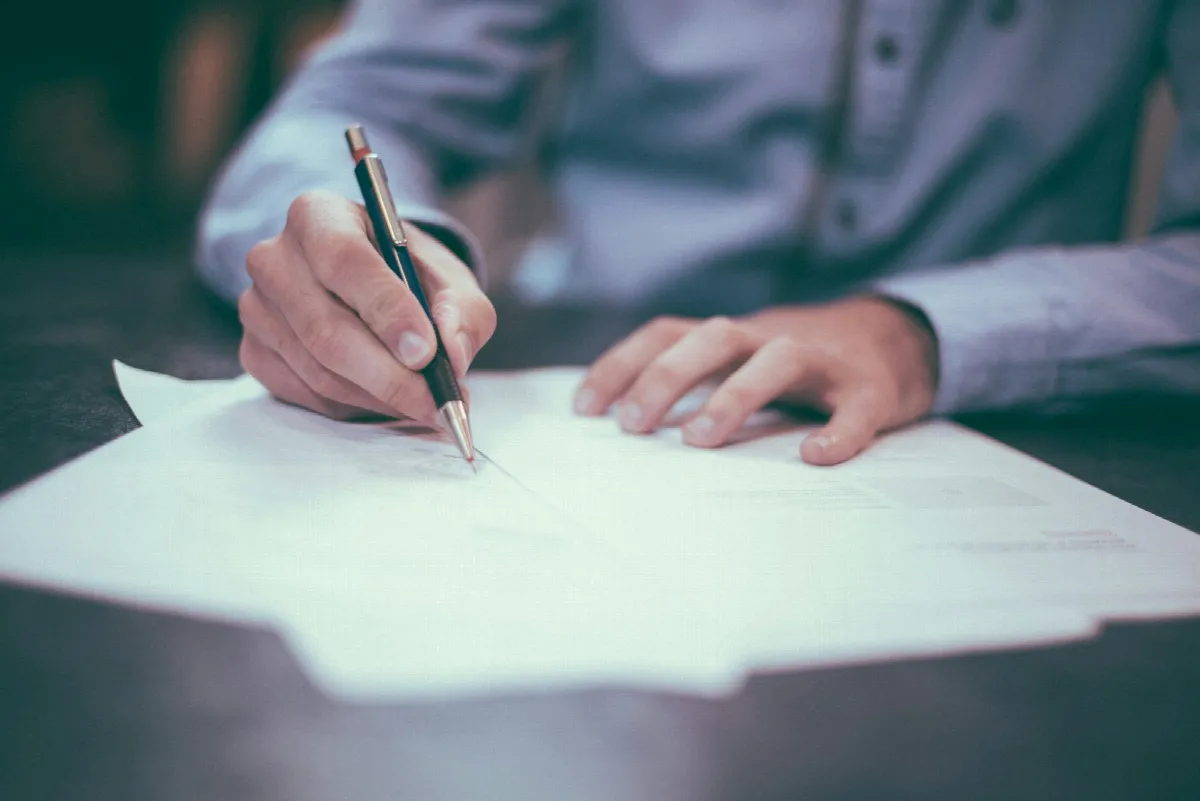 A man in a dress shirt signing a printed letter at a desk with a pen