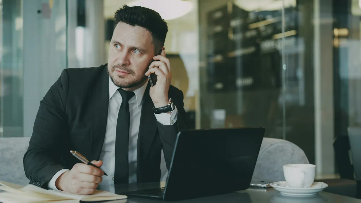A loan broker in a dark suit takes a phone call at a cafe table, pen in hand and laptop open