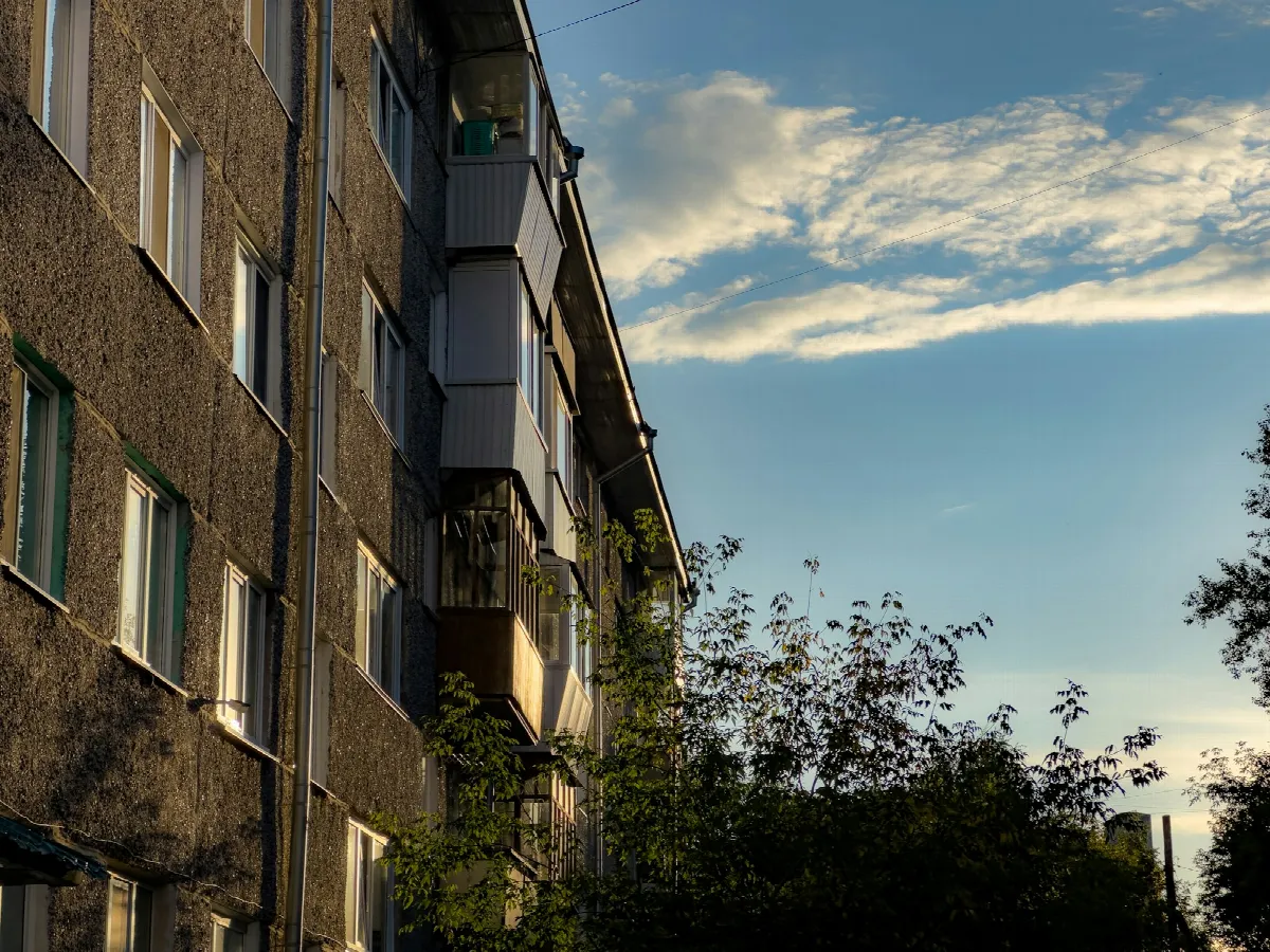 An older brick apartment building with rows of windows and small balconies, trees and a blue evening sky beside it