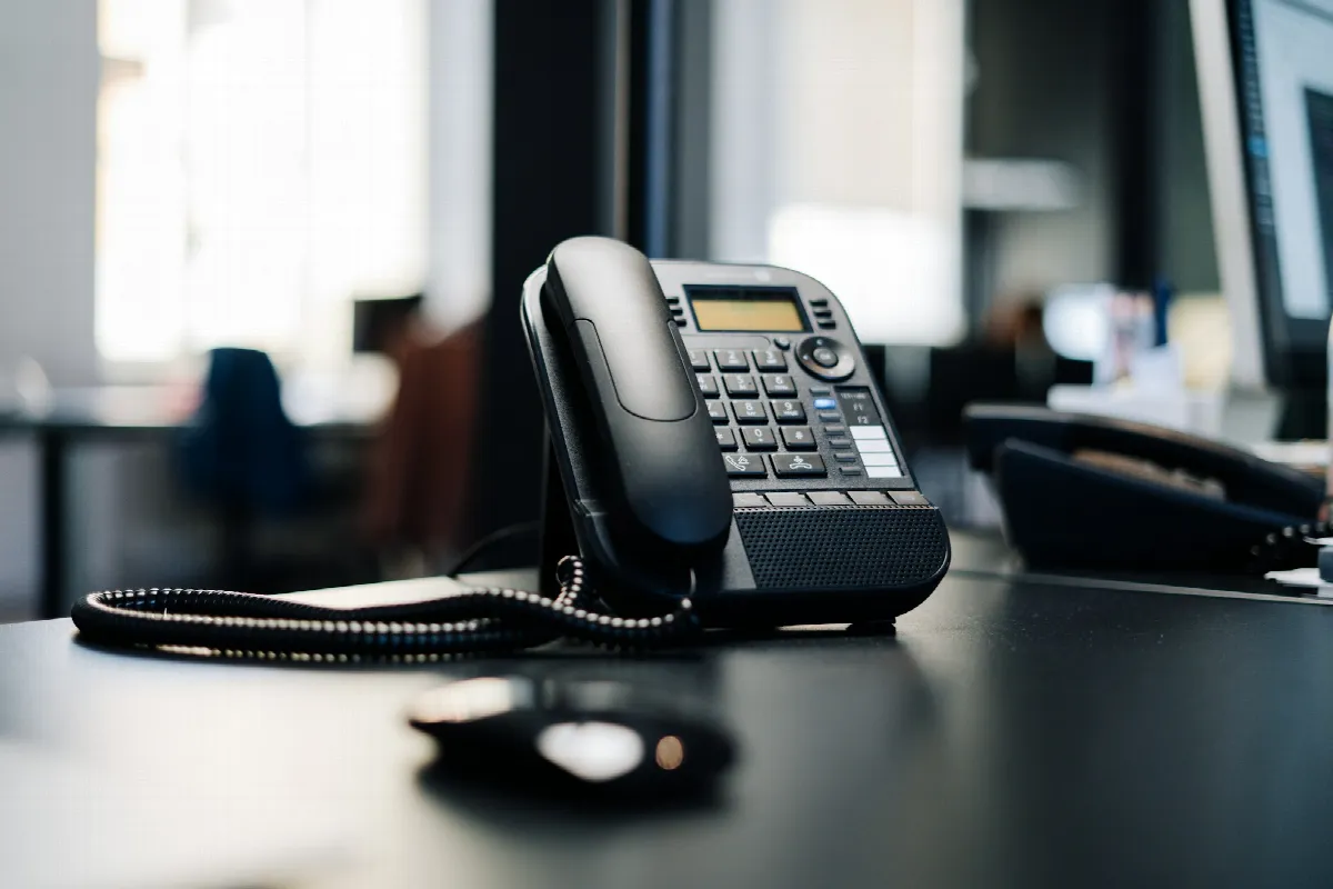 A black IP desk phone with a handset and display sitting on a dark wooden table