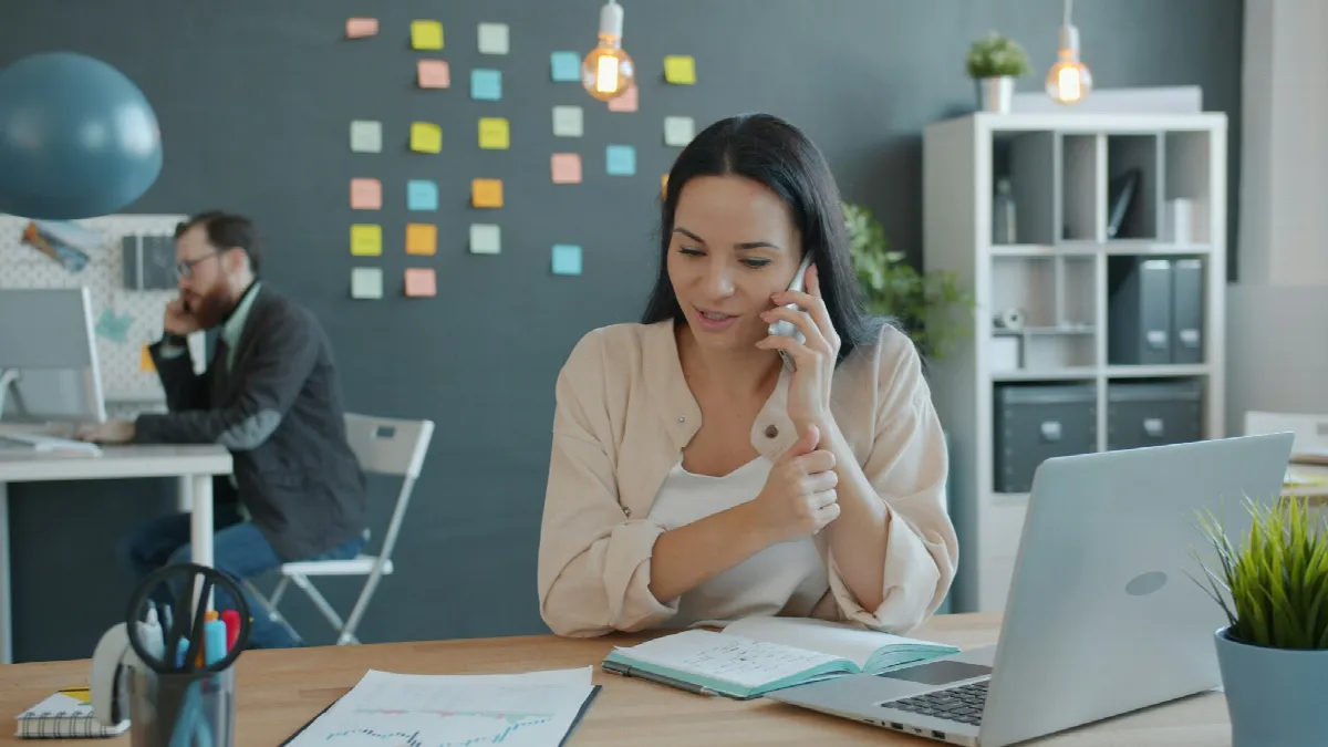 A woman talks on the phone at her desk with a laptop open in front of her