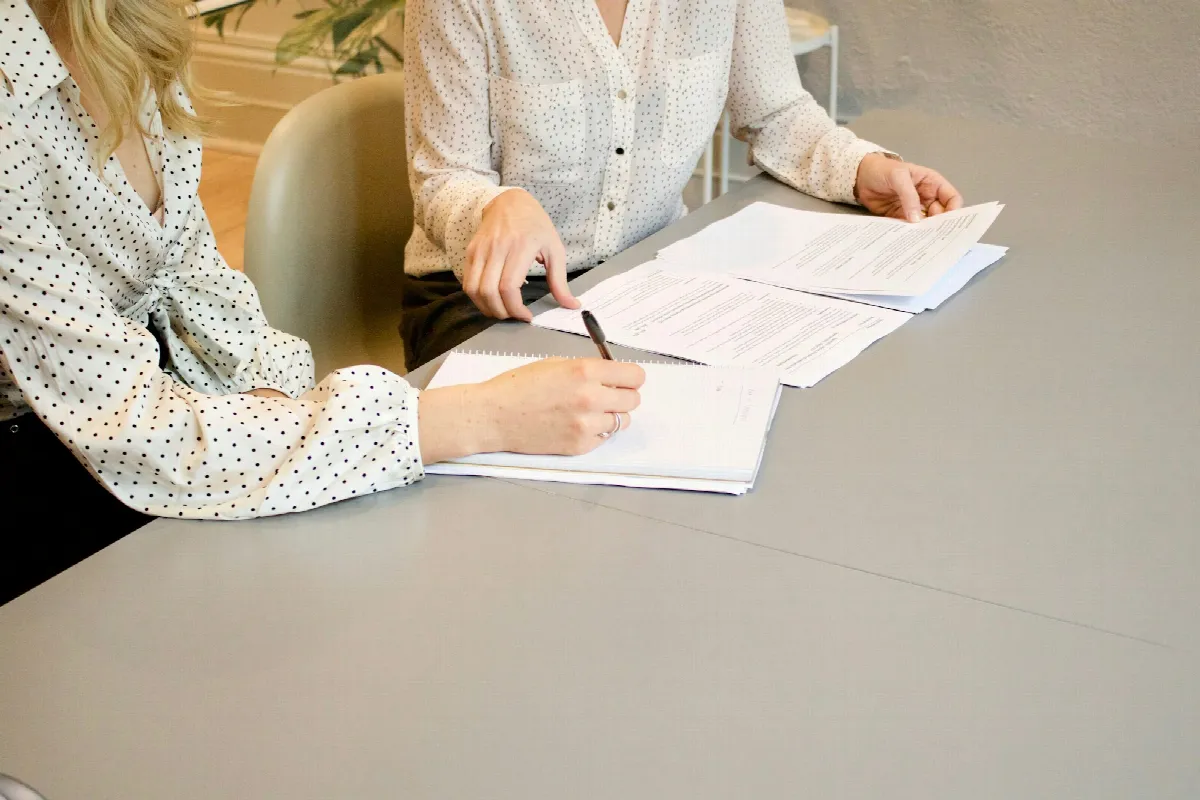 Two women at a gray table, one signing a printed form while the other holds the next page