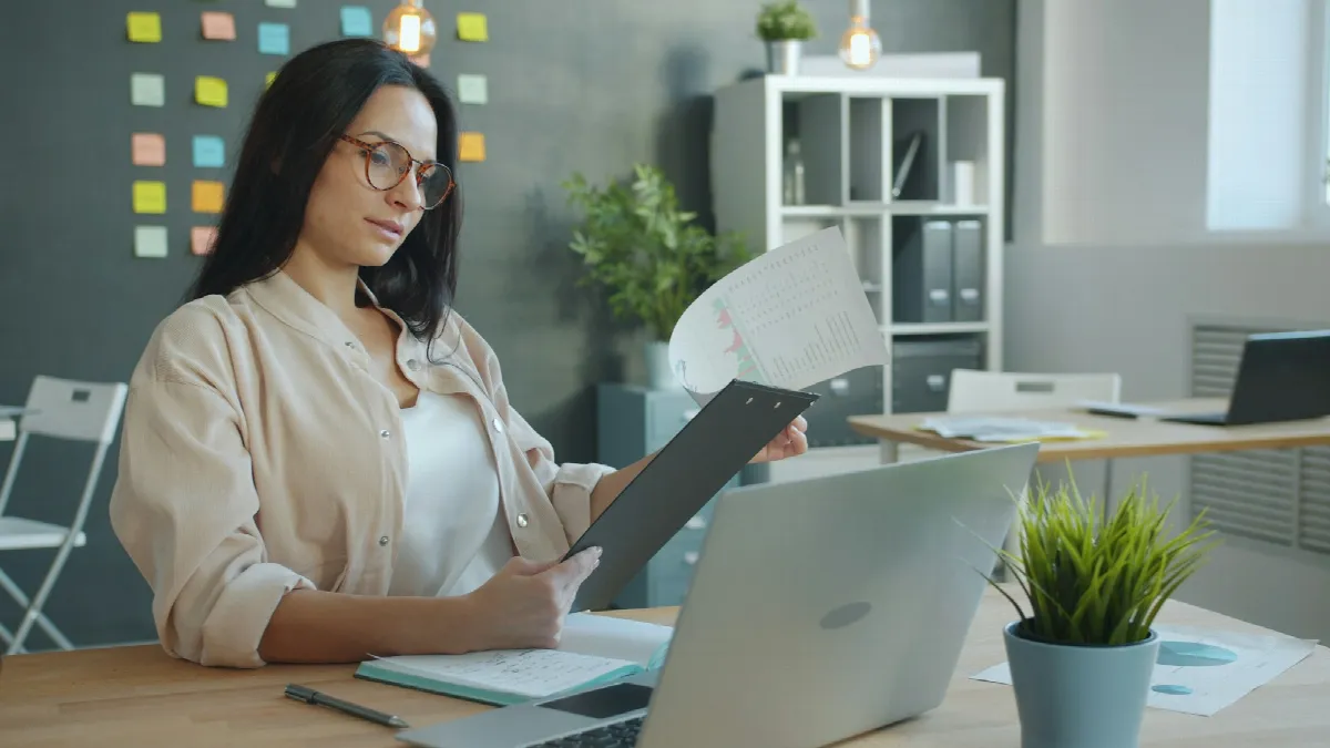 A woman in glasses holds up a document while reviewing information on her laptop at an office desk