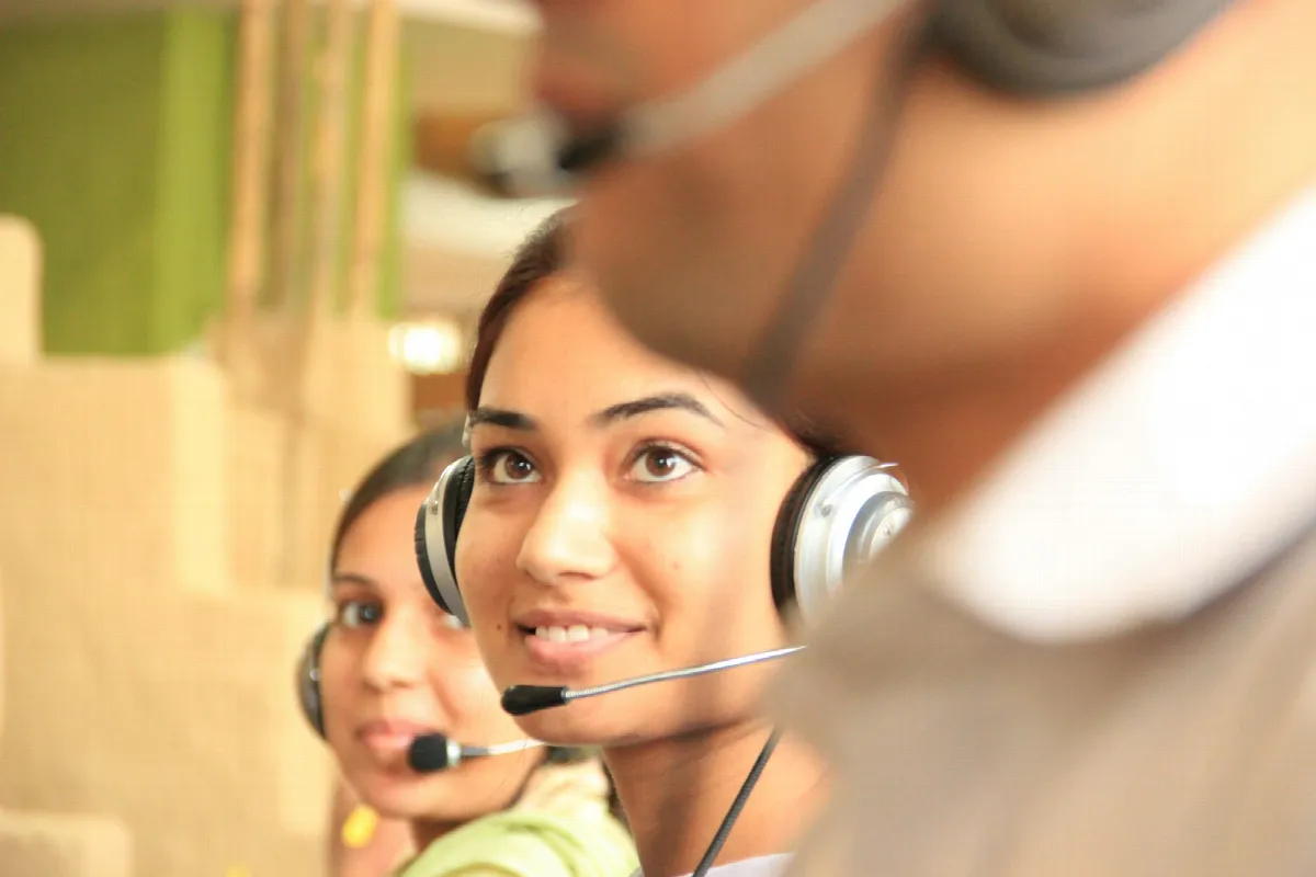Two women wearing headsets work at computers in a bright office