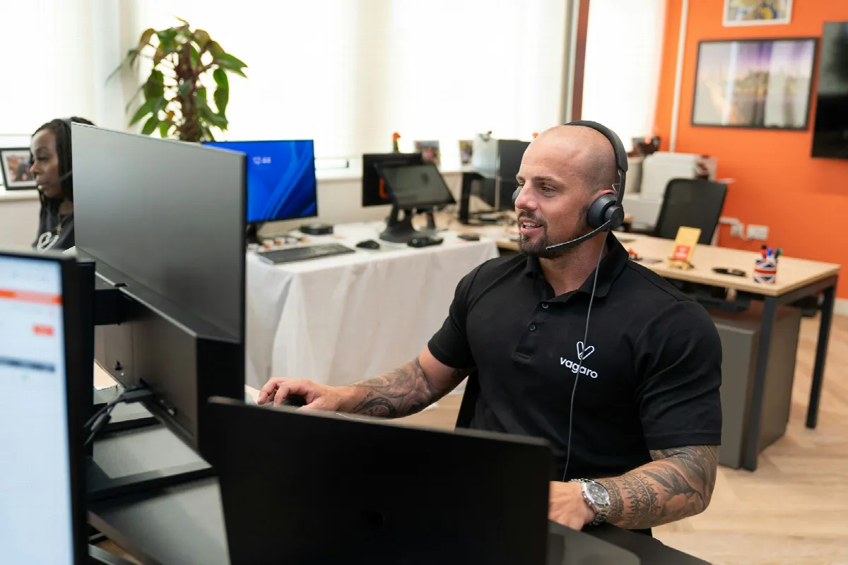 A man wearing a headset sits in front of a computer, ready to take calls