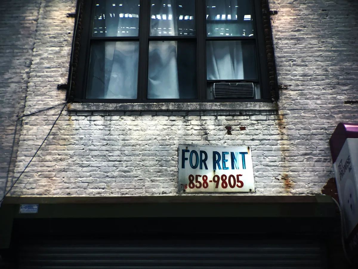 A worn For Rent sign with a phone number painted on it hangs on a brick wall below an apartment window