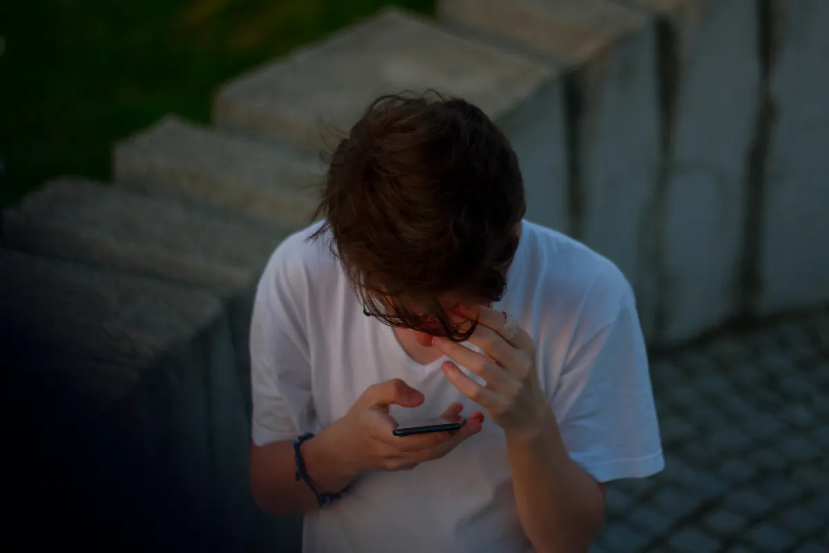 A young man in a white T-shirt checks his smartphone outdoors at dusk, about to run a reverse phone lookup on a missed call
