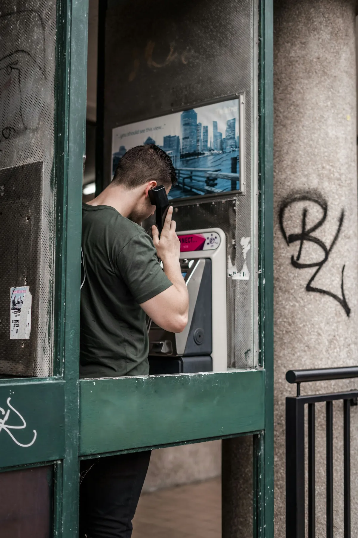 A man in a green T-shirt makes a call from a public payphone booth with graffiti on the wall