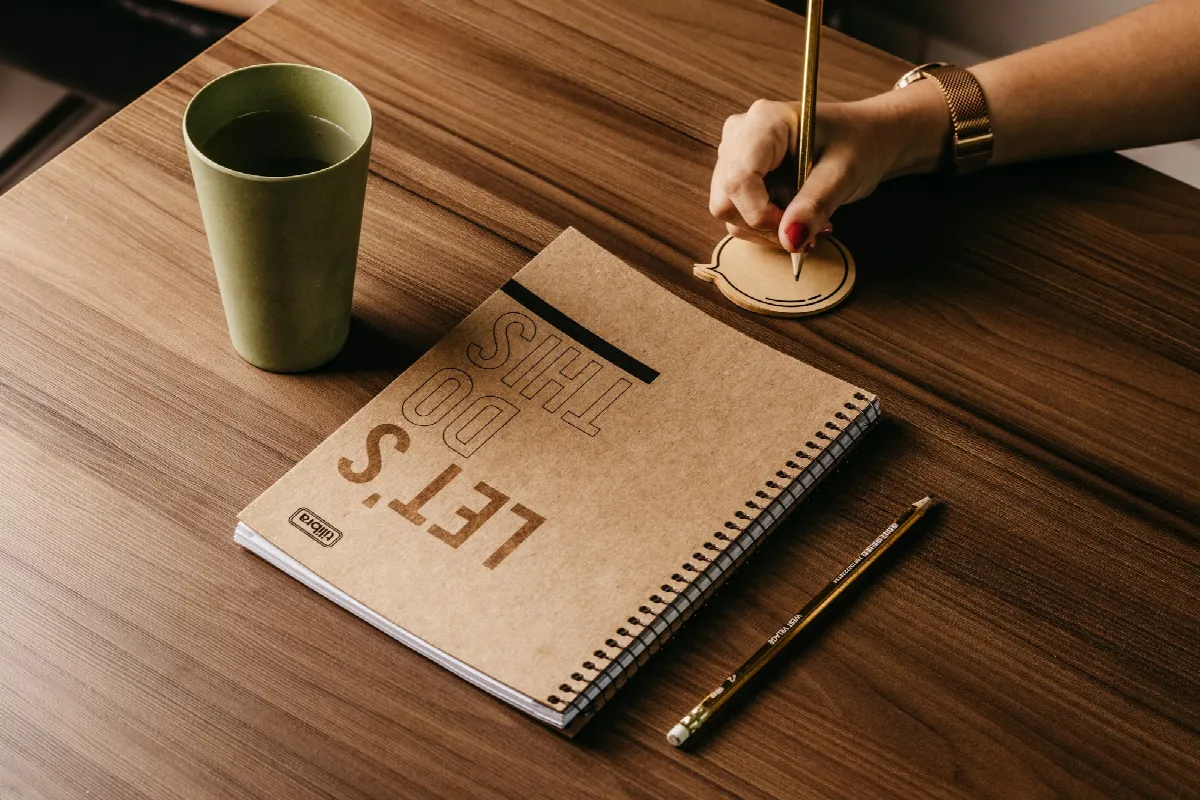 A hand holding a pen over a cardboard notebook on a wooden table beside a green cup