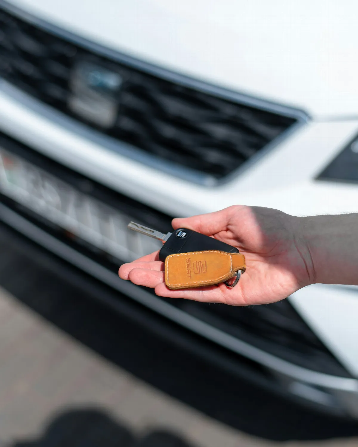A hand holding out a car key with a tan leather fob in front of the grille of a white car