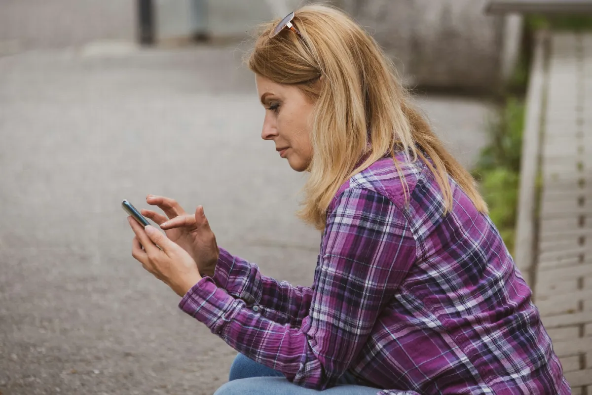 A woman in a plaid shirt sitting outdoors and frowning slightly at a message on her phone