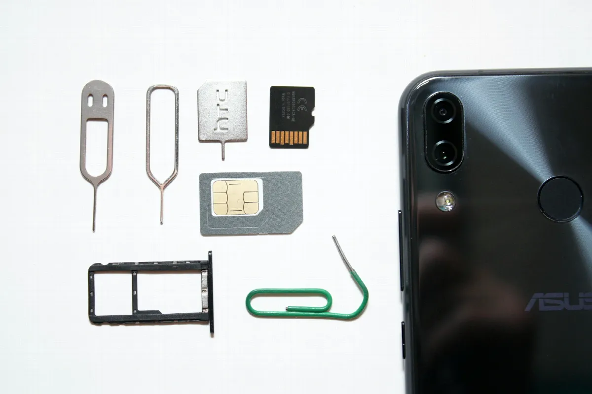 A smartphone beside a removed SIM card, an empty SIM tray, a memory card and two ejector pins on a white surface