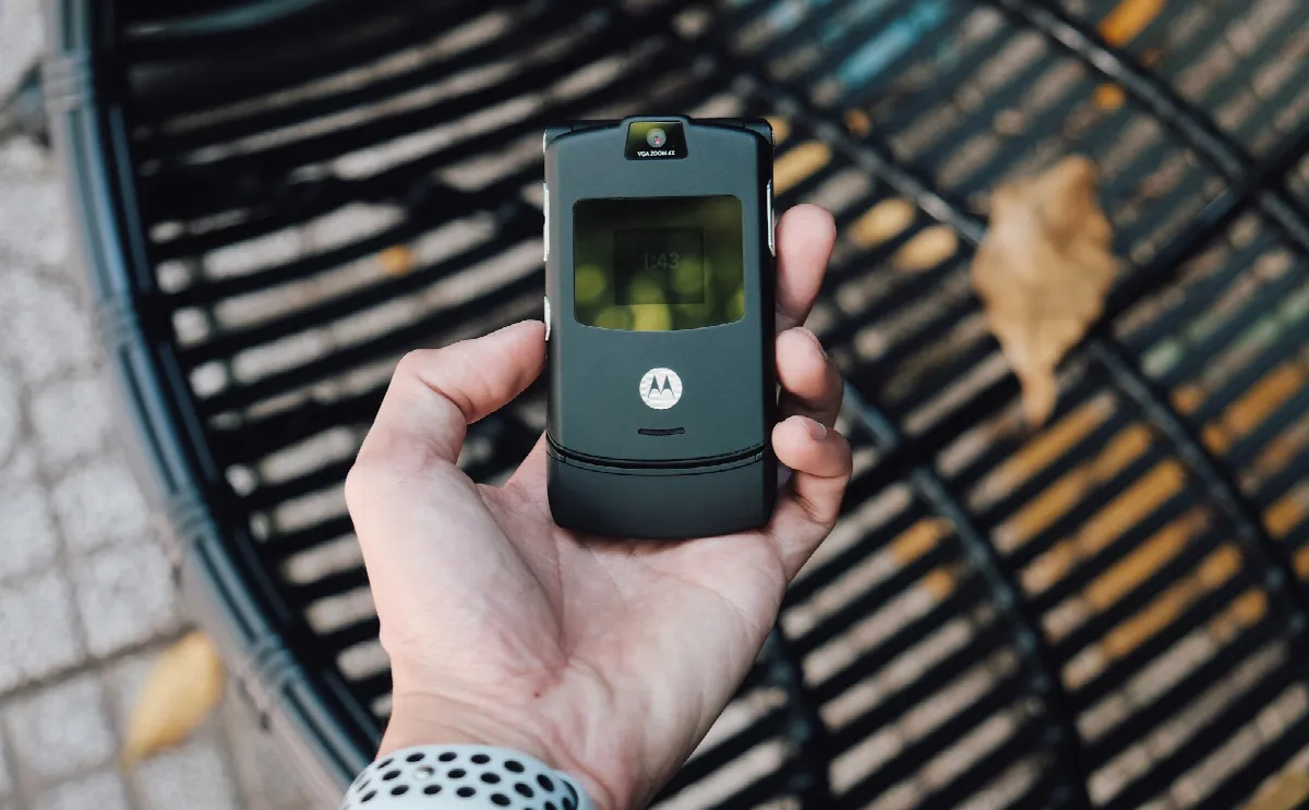 A hand holding a slim black flip phone above a round metal grate outdoors