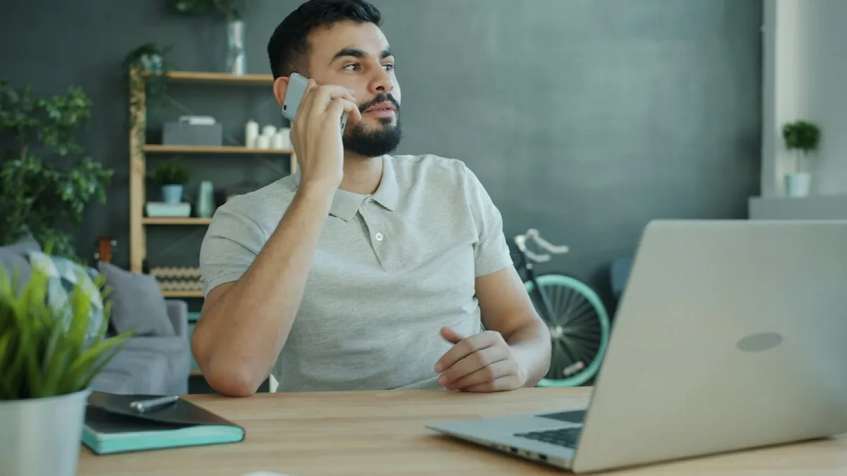 A man at a desk with a laptop talking on his phone while looking off to the side