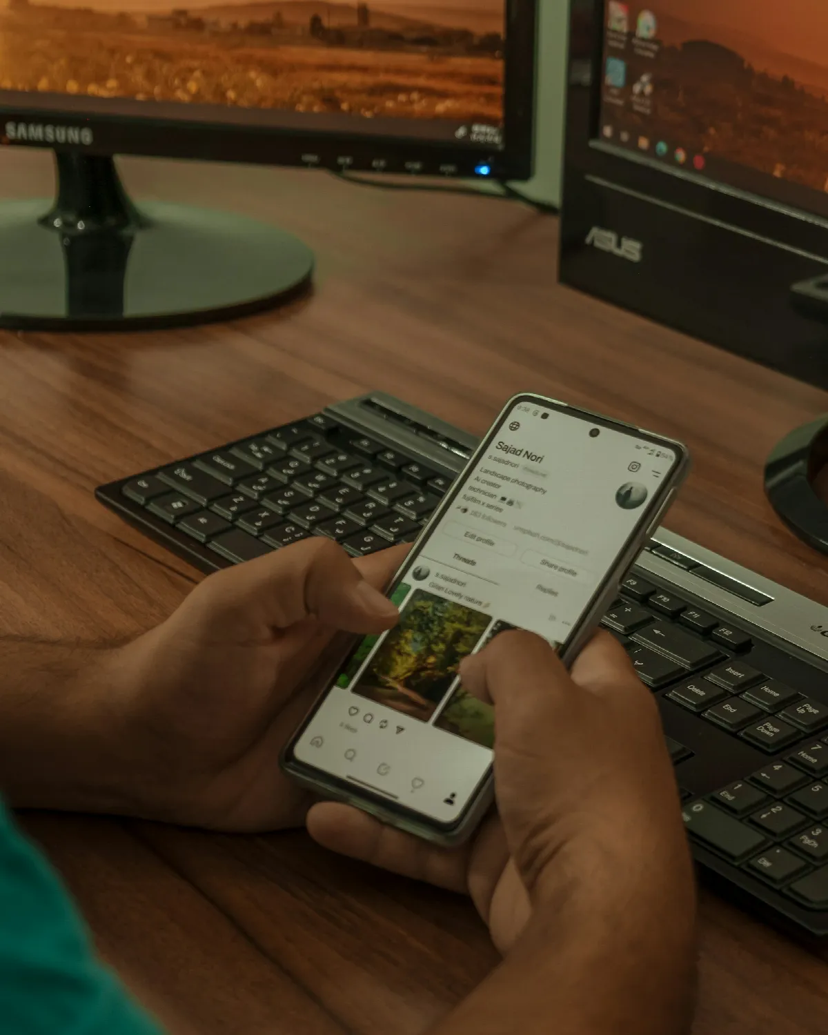 A person holding a smartphone over a keyboard at a desk with two monitors