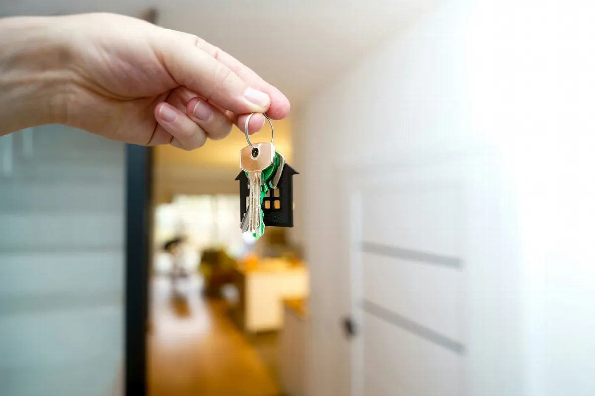 A hand holds up a single house key on a small house-shaped key ring inside a bright apartment hallway