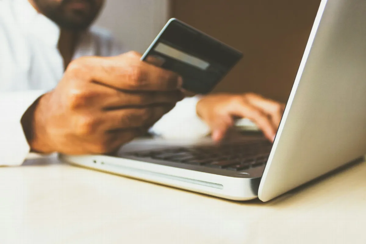 A man holds a payment card in one hand while typing on a laptop at a white desk