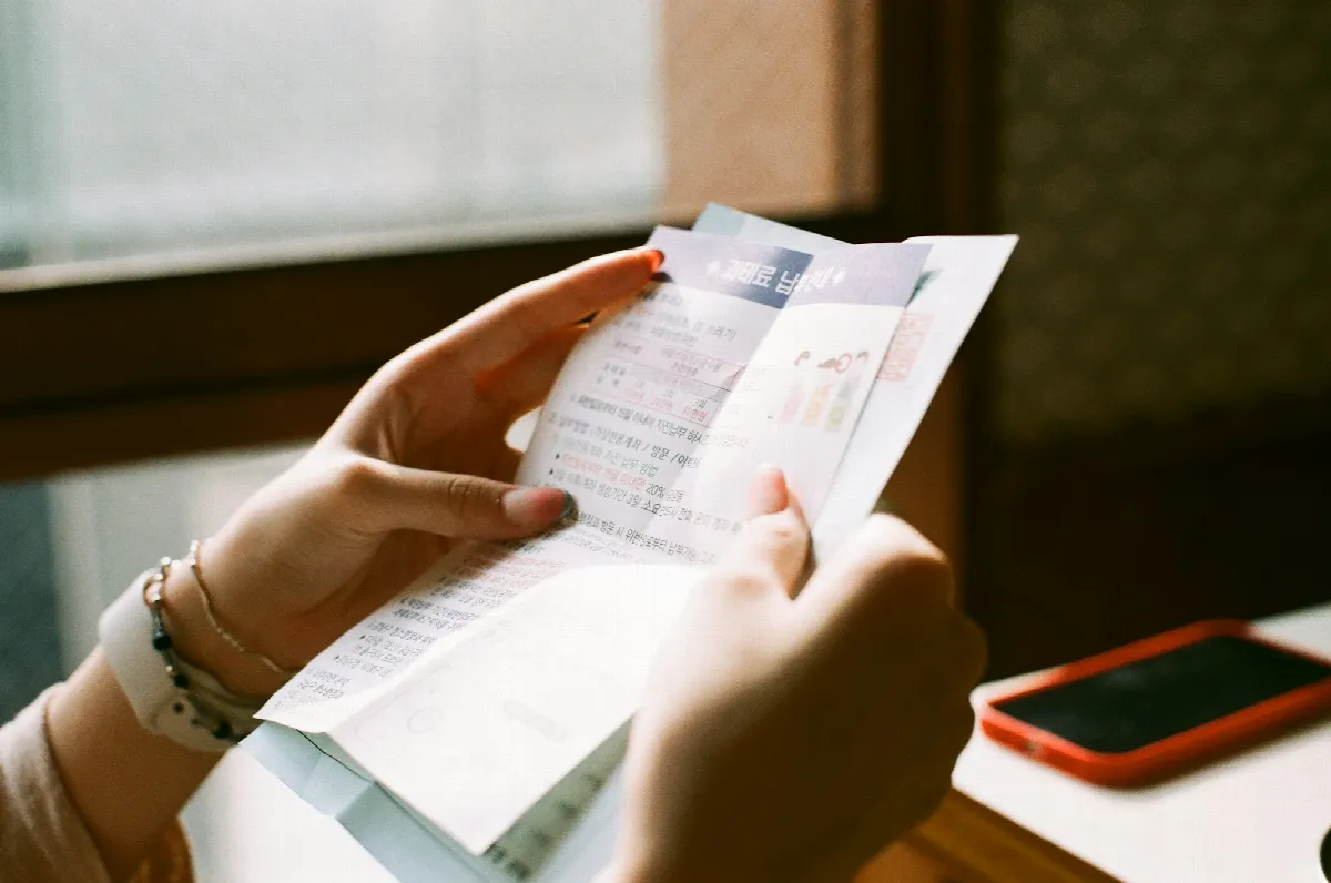 A person holds open a folded paper letter with printed text, a phone with a red case on the table beside them