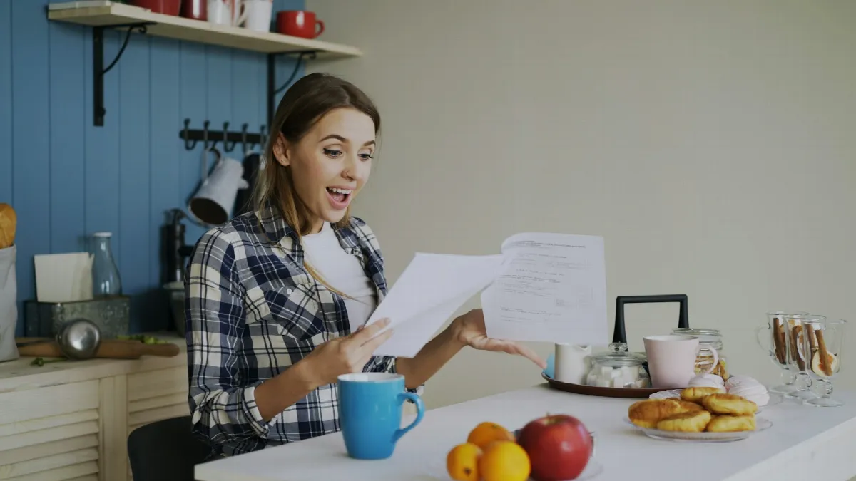 A woman at a kitchen table reacting with surprise as she reads a printed letter