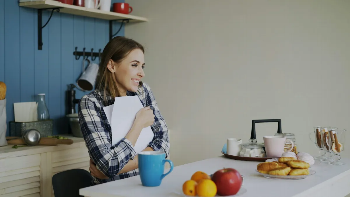 A young woman at a kitchen table reading a letter over breakfast, smiling at what it says