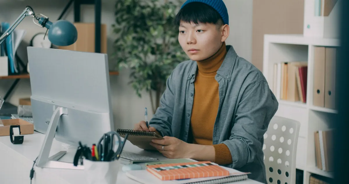 A person in a knit cap and orange turtleneck taking notes at a desk while reading a computer screen