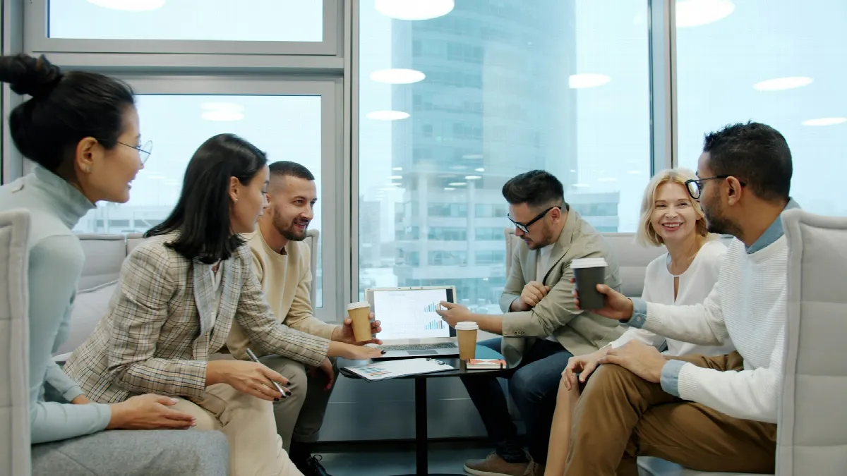 A group of coworkers with coffee cups sitting around a laptop on a low table in an office with tall windows