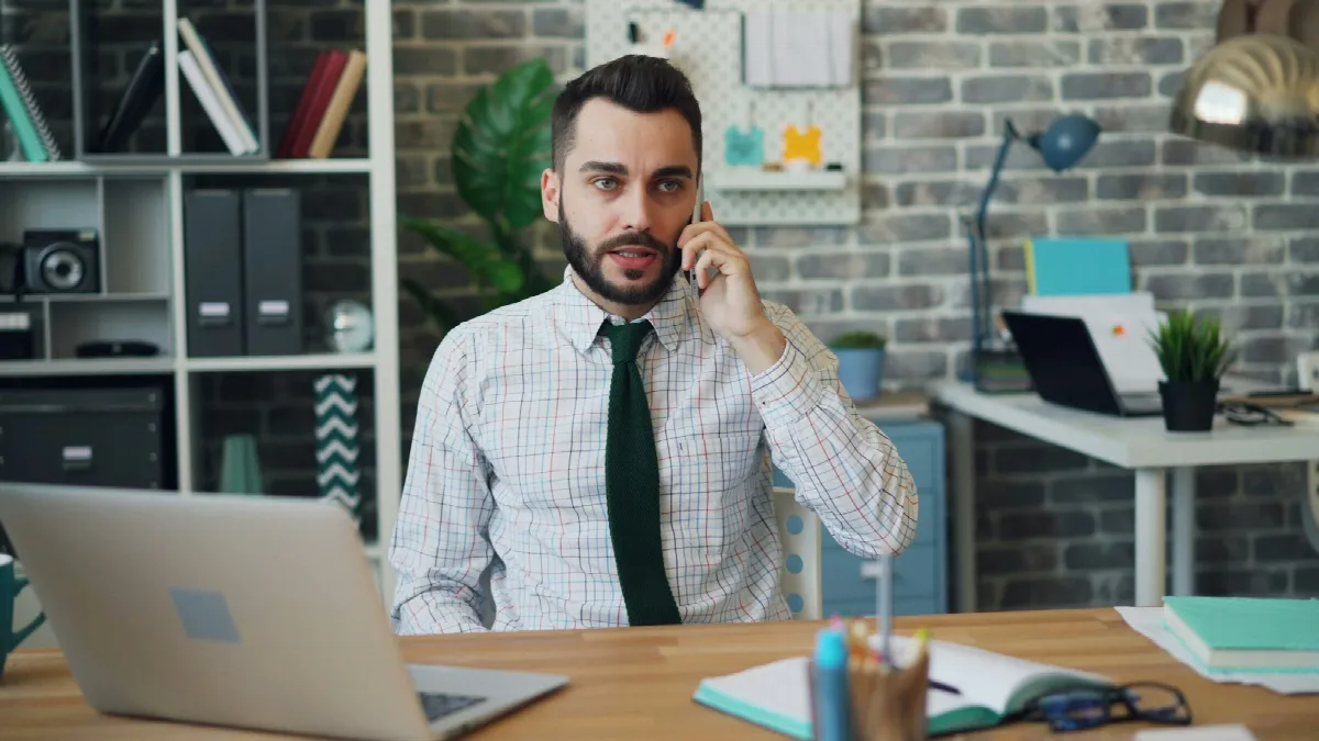 A loan rep in a shirt and tie talks on his phone at a desk, an open laptop and a notebook in front of him