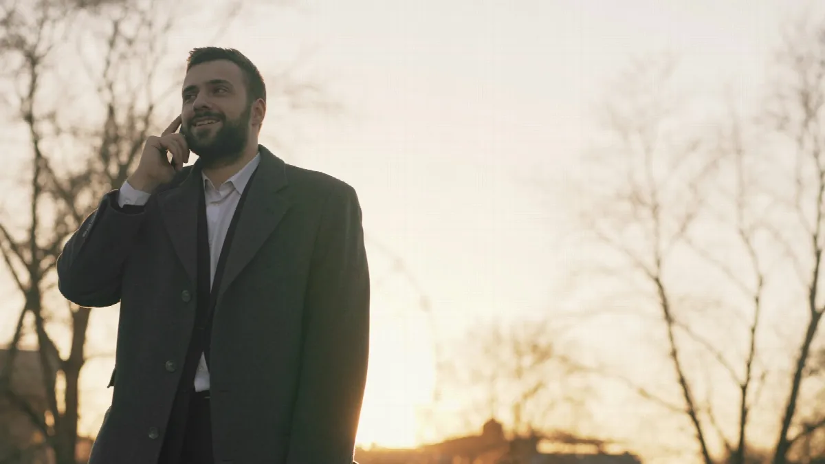 A smiling man in a long coat talks on his phone outdoors at sunset, the kind of caller an ai loan prequalification happens with
