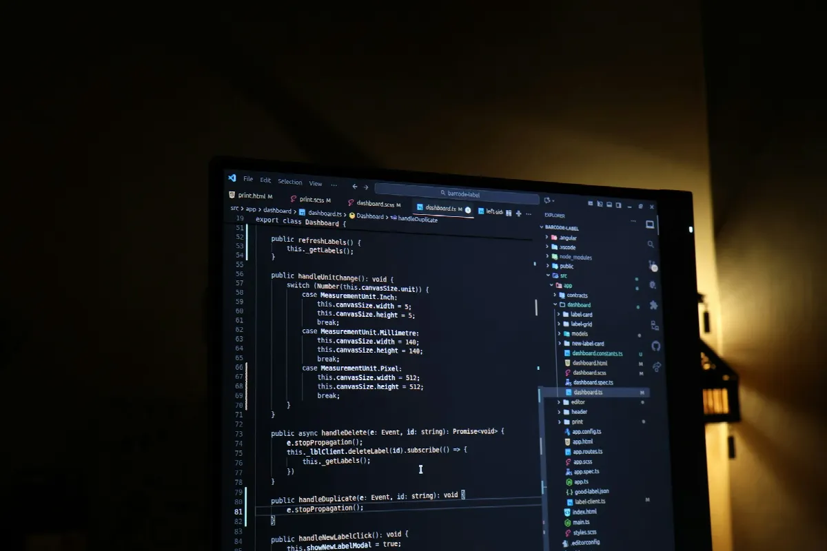A monitor in a dark room showing code in an editor, with a warm lamp glowing beside it