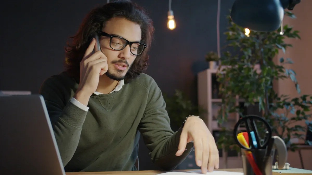 A man in glasses on his phone at an office desk late in the evening, a laptop open in front of him