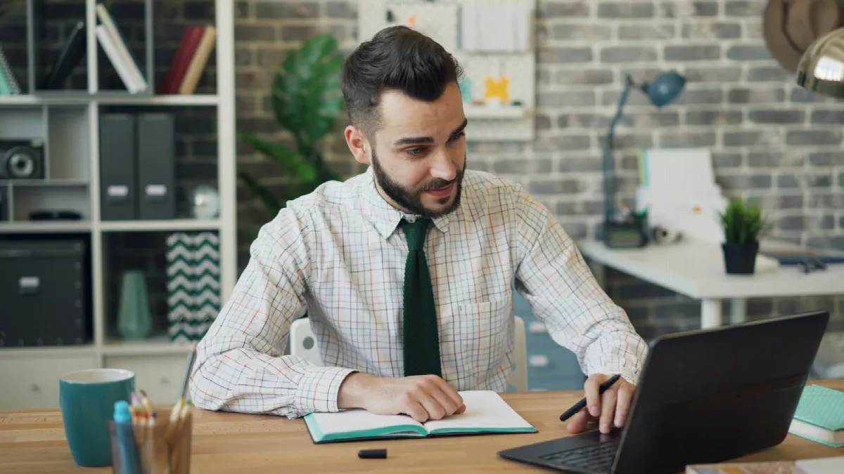 A man in a checked shirt and green tie smiles at a notebook while working on a laptop at a wooden office desk