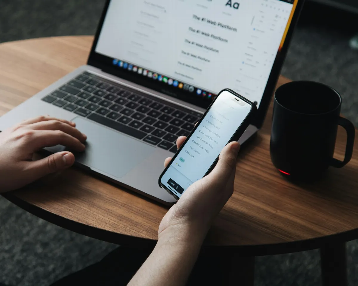 A person holds a smartphone in front of an open laptop on a round wooden table next to a black mug