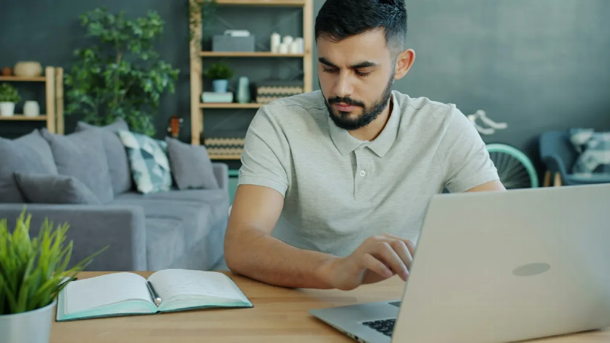 A man in a gray polo shirt types on a laptop at a wooden table in a living room, an open notebook beside him