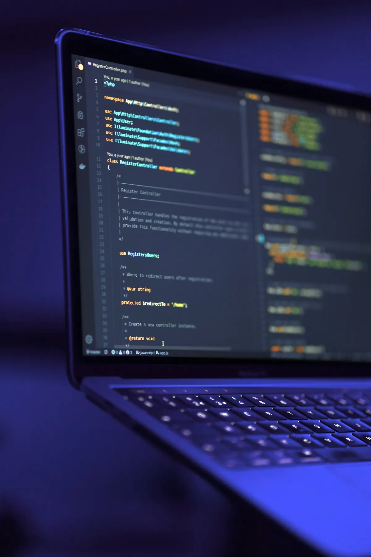 A laptop on a dark desk, its screen full of code in a dark-themed editor, lit in deep blue