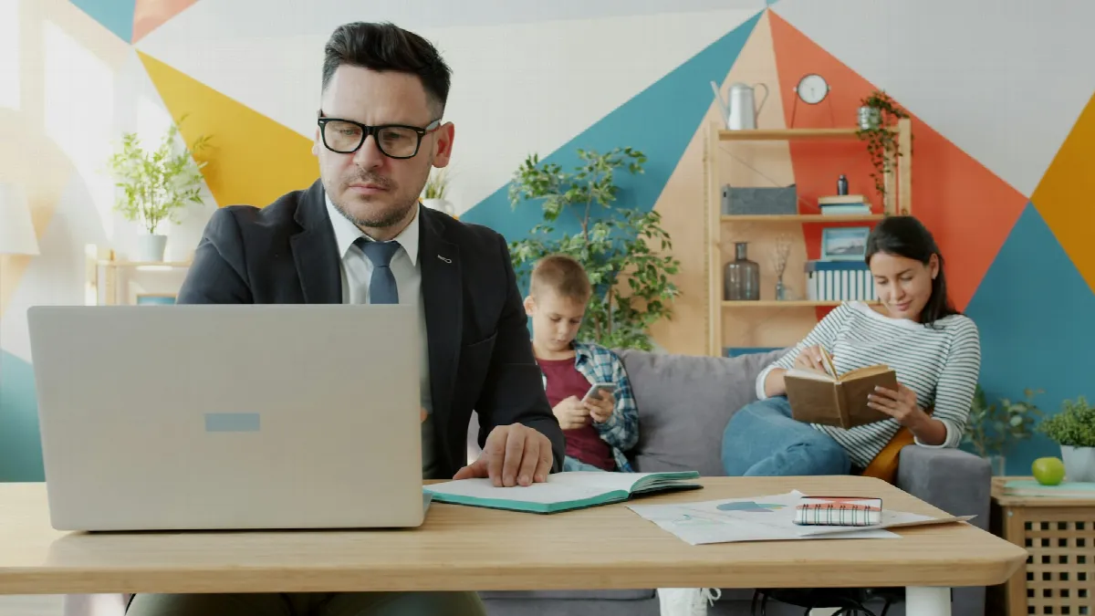 A man in a suit works on a laptop at a table at home while a child and a woman read on the couch behind him